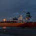 A large oil tanker illuminated at night, docked at an industrial harbor under a dark sky.