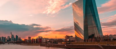 The modern glass tower of the European Central Bank headquarters in Frankfurt reflecting a colorful sunset.
