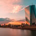 The modern glass tower of the European Central Bank headquarters in Frankfurt reflecting a colorful sunset.