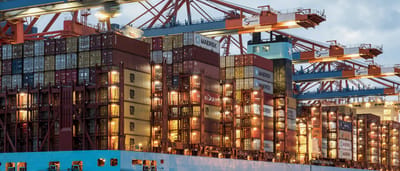 A massive container ship stacked with colorful shipping containers is loaded by illuminated cranes at a bustling harbor at dusk.
