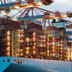 A massive container ship stacked with colorful shipping containers is loaded by illuminated cranes at a bustling harbor at dusk.