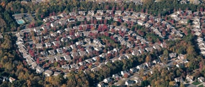 An aerial view of a large suburban neighborhood with identical houses arranged in concentric circles among autumn trees.