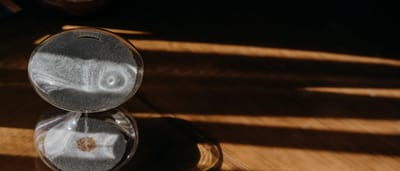A glass sand timer sits on a wooden desk bathed in warm sunlight, casting long shadows.