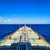 A view looking forward over the expansive deck of a large shipping tanker sailing across the open ocean under a bright blue sky.