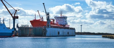 A large orange ship sits in a floating dry dock at an industrial shipyard under a bright blue sky.