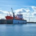 A large orange ship sits in a floating dry dock at an industrial shipyard under a bright blue sky.