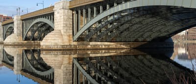 A solid stone and steel bridge stretches across a calm river under a blue sky, its arches reflecting in the water.