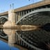 A solid stone and steel bridge stretches across a calm river under a blue sky, its arches reflecting in the water.
