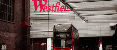 A red double-decker bus drives past the large red Westfield shopping center sign in London.