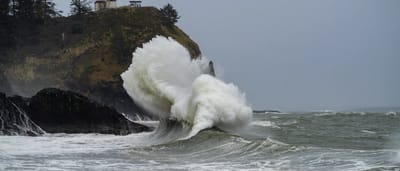 A massive white ocean wave crashes violently against a dark rocky cliffside beneath a standing lighthouse.