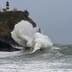 A massive white ocean wave crashes violently against a dark rocky cliffside beneath a standing lighthouse.
