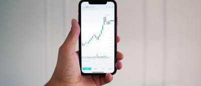 A hand holding a smartphone displaying a financial candlestick chart on a trading application.