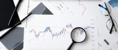 A top-down view of printed stock market charts, a magnifying glass, eyeglasses, and pens arranged on a desk.