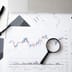 A top-down view of printed stock market charts, a magnifying glass, eyeglasses, and pens arranged on a desk.