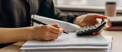 A person's hands using a calculator while reviewing a stack of financial documents on a wooden desk.