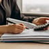 A person's hands using a calculator while reviewing a stack of financial documents on a wooden desk.