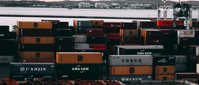 Stacks of cargo shipping containers fill a bustling industrial port at twilight.