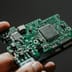 A close-up view of a person's hand holding a green semiconductor circuit board with microchips.