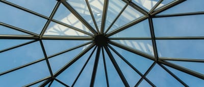 A symmetrical view from below of a modern, geometric glass ceiling against a clear blue sky.
