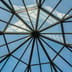 A symmetrical view from below of a modern, geometric glass ceiling against a clear blue sky.