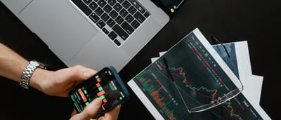 An overhead view of a desk featuring a laptop, printed candlestick charts, and a person holding a smartphone displaying a trading app.