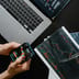 An overhead view of a desk featuring a laptop, printed candlestick charts, and a person holding a smartphone displaying a trading app.