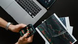 An overhead view of a desk featuring a laptop, printed candlestick charts, and a person holding a smartphone displaying a trading app.