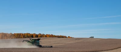 A green combine harvester kicks up dust as it works through a large, rolling field of soybeans under a clear blue sky.