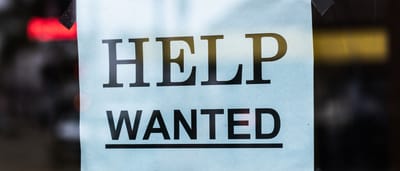 A printed Help Wanted sign taped to the glass window of a storefront.