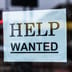 A printed Help Wanted sign taped to the glass window of a storefront.