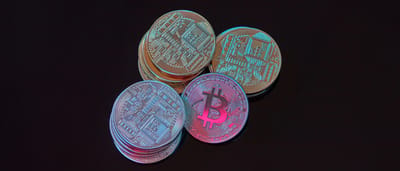 A stack of physical gold Bitcoin tokens sitting in heavy shadow against a pitch-black background.