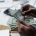 Close-up of hands counting twenty-dollar bills over financial documents and a smartphone on a desk.
