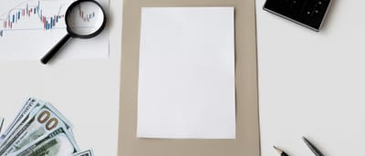 A blank white document sits on a desk surrounded by a stock chart, a magnifying glass, a calculator, and hundred dollar bills.