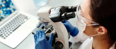 A scientist wearing blue gloves examines a sample under a microscope next to a laptop displaying biological data.