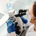 A scientist wearing blue gloves examines a sample under a microscope next to a laptop displaying biological data.
