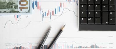 A top-down view of printed financial charts, a calculator, two pens, and a portion of a U.S. hundred-dollar bill on a desk.