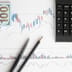 A top-down view of printed financial charts, a calculator, two pens, and a portion of a U.S. hundred-dollar bill on a desk.