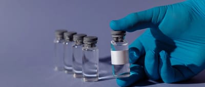 A blue-gloved medical professional holds up a small glass vial with a blank white label against a soft purple background.