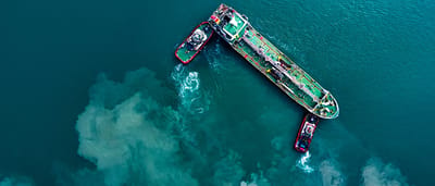 An aerial top-down view of an oil tanker being guided by tugboats through deep teal waters.