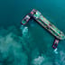 An aerial top-down view of an oil tanker being guided by tugboats through deep teal waters.