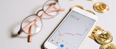 A smartphone displaying an upward-trending cryptocurrency chart lies next to gold Bitcoin tokens and stylish eyeglasses on a light surface.