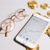 A smartphone displaying an upward-trending cryptocurrency chart lies next to gold Bitcoin tokens and stylish eyeglasses on a light surface.