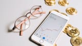 A smartphone displaying an upward-trending cryptocurrency chart lies next to gold Bitcoin tokens and stylish eyeglasses on a light surface.