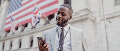 A smiling businessman checks his smartphone outside the New York Stock Exchange with a large American flag in the background.