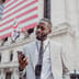 A smiling businessman checks his smartphone outside the New York Stock Exchange with a large American flag in the background.