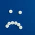 White round pills arranged in the shape of a frowning sad face on a solid blue background.