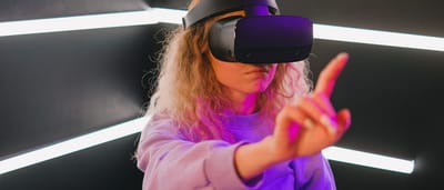 A young woman wearing a virtual reality headset reaches out to interact with a digital environment illuminated by pink and blue neon lights.