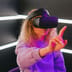 A young woman wearing a virtual reality headset reaches out to interact with a digital environment illuminated by pink and blue neon lights.