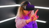 A young woman wearing a virtual reality headset reaches out to interact with a digital environment illuminated by pink and blue neon lights.
