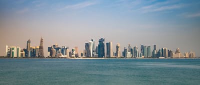 A bright, panoramic view of the modern Doha skyline rising across the water under a clear blue sky.